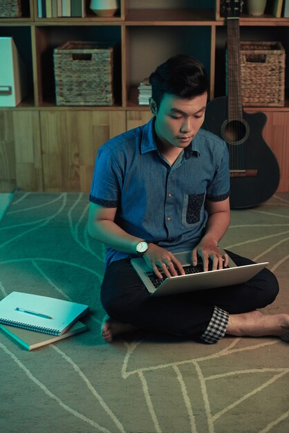 Jeune Homme Asiatique Concentré En Montre-bracelet Assis Avec Les Jambes Croisées Sur Un Tapis Et Travaillant Avec Un Ordinateur Portable En...