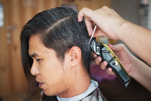 Jeune homme asiatique coupé des cheveux longs aux cheveux courts avec une tondeuse électrique par un coiffeur professionnel en salon de coiffure.