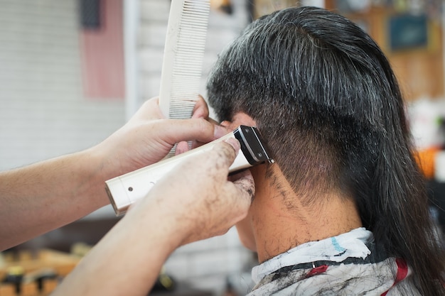 Jeune homme asiatique coupé des cheveux longs aux cheveux courts avec une tondeuse électrique par un coiffeur professionnel en salon de coiffure.