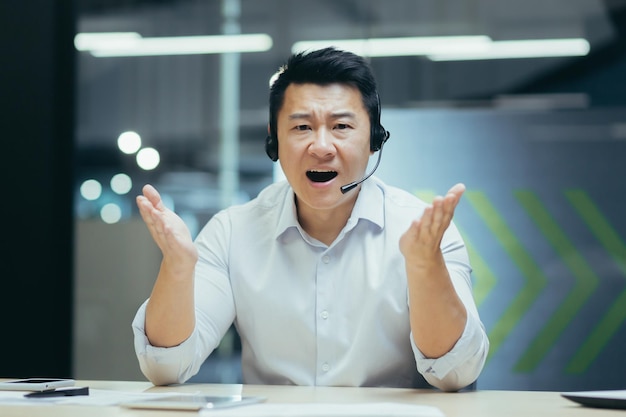 Un jeune homme asiatique est assis au bureau à la table dans un casque et parle avec émotion à