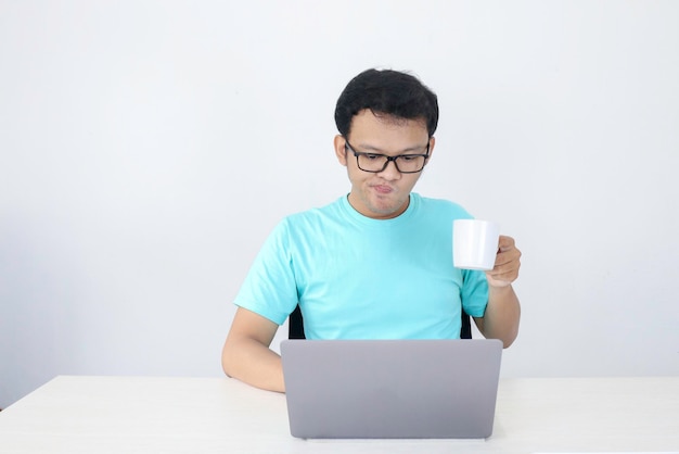 Jeune homme asiatique est sérieux lorsqu'il travaille dans un ordinateur portable avec un verre de café blanc Indonésie Homme porte une chemise bleue Fond gris isolé