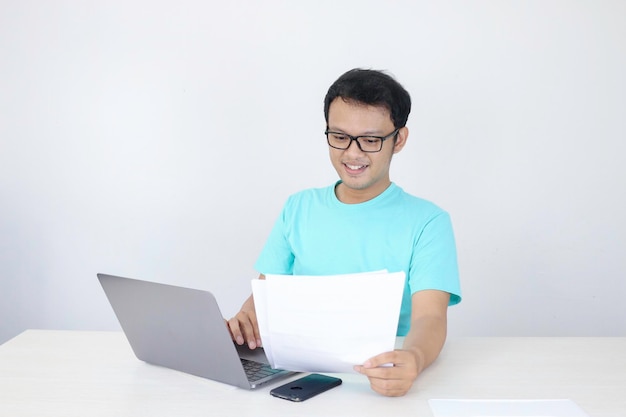 Jeune homme asiatique est souriant et heureux lorsqu'il travaille sur un ordinateur portable et un document à portée de main Homme indonésien portant une chemise bleue