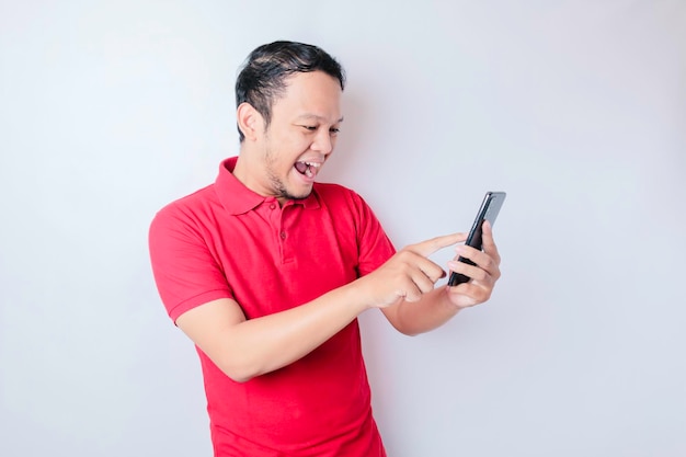 Un jeune homme asiatique avec une expression heureuse et réussie portant une chemise rouge et tenant un smartphone isolé sur fond blanc