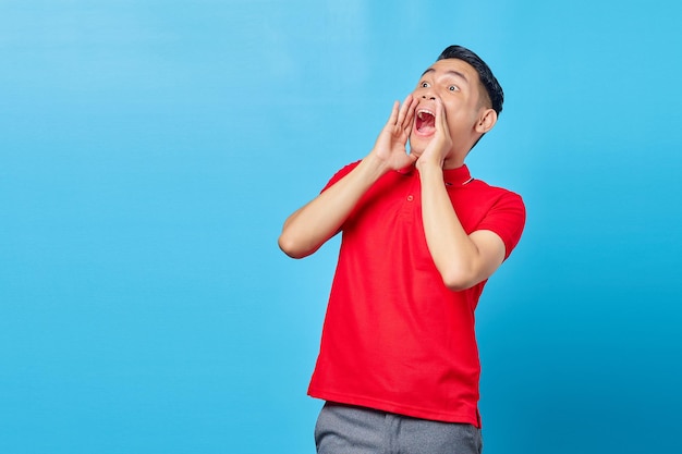 Jeune homme asiatique heureux annonçant des informations avec une tasse de mains autour de la bouche isolée sur fond de studio bleu