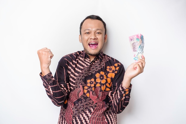 Un jeune homme asiatique heureux avec une expression réussie portant une chemise batik et tenant de l'argent en roupie indonésienne isolé sur fond blanc