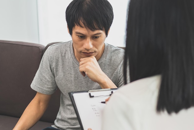 Jeune homme asiatique, un homme a des symptômes mentaux qui doivent avoir été une thérapie et du stress, assis sur un canapé pour consulter un psychologue pendant la séance en prenant des notes pour savoir comment traiter le thérapeute.