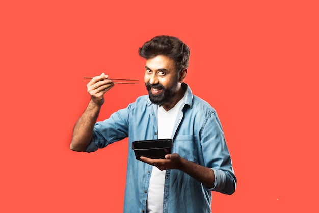 Jeune homme asiatique indien mangeant des plats à emporter dans la boîte à l'aide d'une cuillère ou de baguettes, debout sur le rouge
