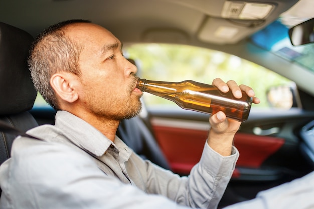 Jeune homme asiatique ivre conduit une voiture avec une bouteille de bière avec fond de coucher de soleil