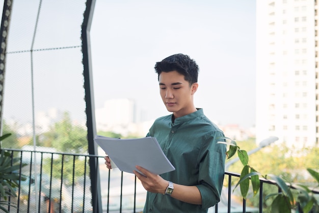 Jeune homme asiatique lisant des documents, debout dans un balcon extérieur