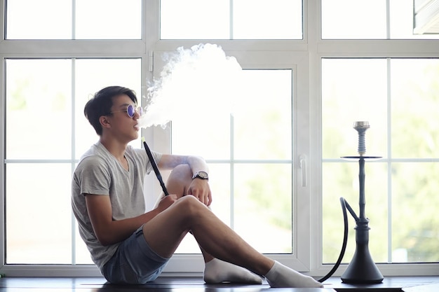 Jeune homme asiatique à lunettes de soleil fumant un narguilé à la fenêtre