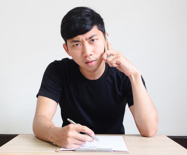 Jeune homme asiatique parlant avec le visage sérieux de téléphone tenant le stylo et le presse-papiers sur le bureau