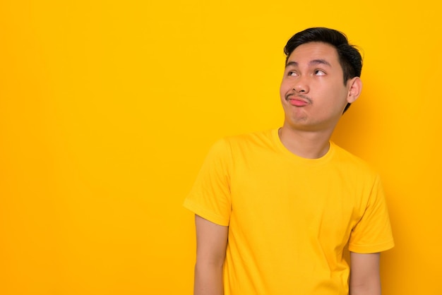 Jeune homme asiatique pensif en t-shirt décontracté regardant l'espace de copie en pensant à quelque chose d'isolé sur fond jaune Concept de style de vie des gens