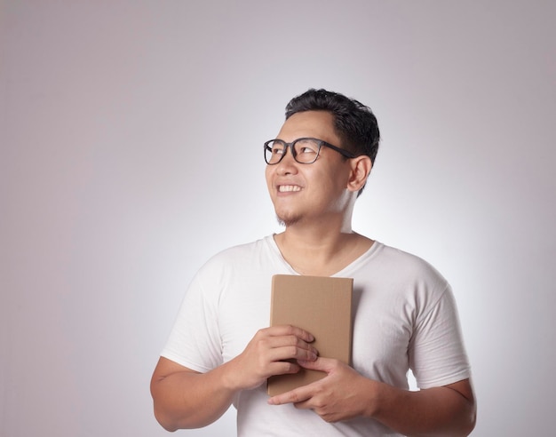 Jeune homme asiatique portant une chemise blanche décontractée tenant un livre avec un geste pensant et un sourire