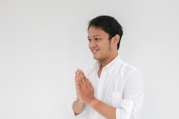 Jeune homme asiatique portant une chemise blanche salue les mains avec un grand sourire sur son visage Homme indonésien sur fond gris Eid Mubarak Eid Fitr Eid AlFitr salutation