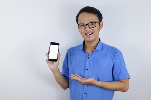 Jeune homme asiatique portant une chemise bleue est debout et souriant pointant sur un espace blanc sur l'écran du smartphone sur fond blanc