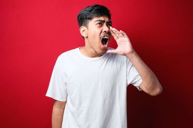 Jeune homme asiatique portant un style t-shirt blanc criant et criant fort à côté avec la main sur la bouche sur fond rouge. notion de communication.