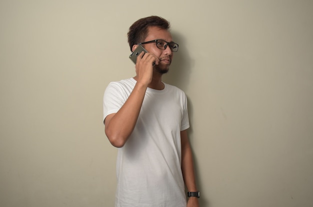Jeune homme asiatique portant un t-shirt blanc debout avec le sourire sur le visage parlant au téléphone