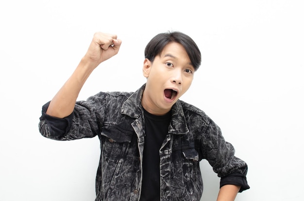 Jeune homme asiatique portant une veste en jean surpris, étonné, wow choqué en levant les bras oui geste
