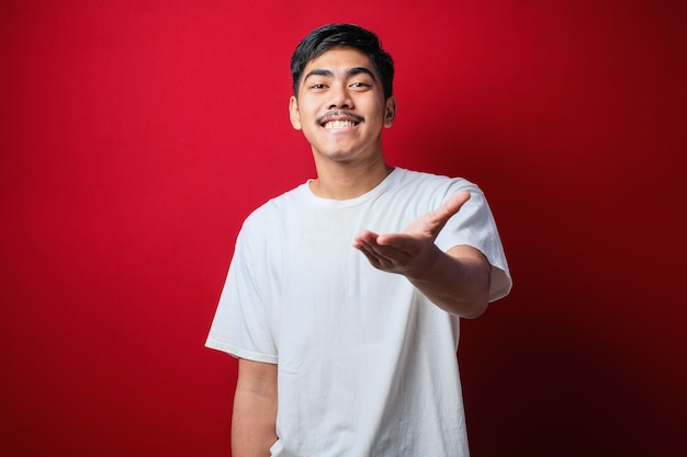 Jeune homme asiatique portant des vêtements blancs souriant amical offrant une poignée de main comme salutation et accueil sur fond rouge. entreprise prospère.