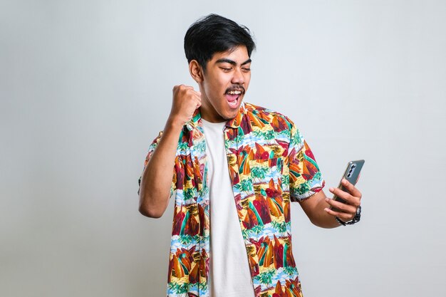 Jeune homme asiatique posant sur fond blanc isolé jouer à des jeux par téléphone faire un geste gagnant.