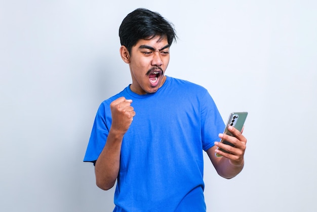 Jeune homme asiatique posant sur fond blanc isolé jouer à des jeux par téléphone faire un geste gagnant.