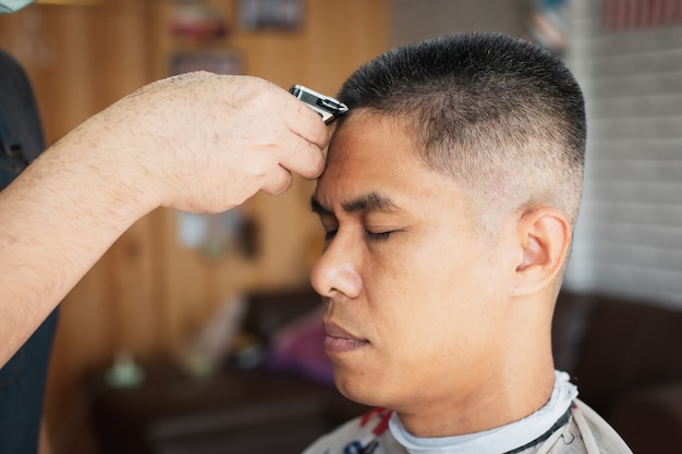 Jeune homme asiatique qui a les cheveux gris se fait couper les cheveux avec une tondeuse électrique par un coiffeur professionnel en salon de coiffure.