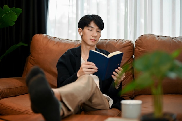 Un jeune homme asiatique qui lit un livre et se détend sur son canapé.