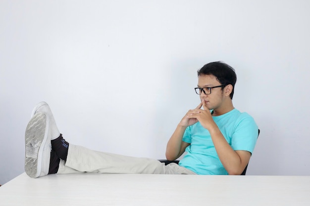 Jeune homme asiatique avec un regard sérieux et concentré avec la jambe sur la table et le bras sur le menton Homme indonésien portant une chemise bleue