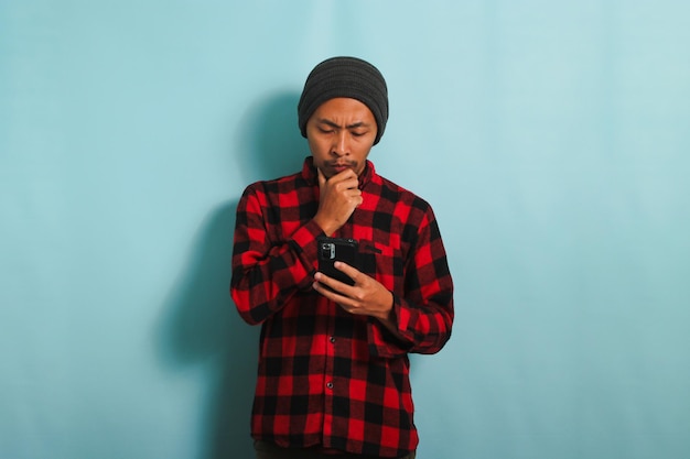 Jeune homme asiatique regarde à travers ses lunettes tout en tenant un téléphone isolé sur fond bleu