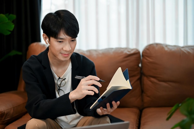 Jeune homme asiatique relaxant qui lit des livres et prend des notes assis sur le canapé.