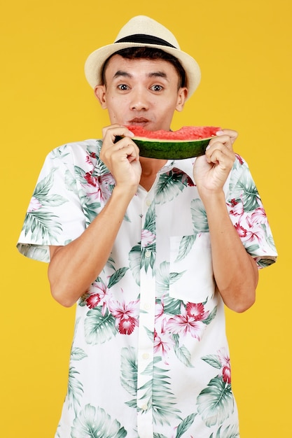 Jeune homme asiatique séduisant en chemise hawaïenne blanche portant un chapeau blanc mangeant une tranche de pastèque avec enthousiasme sur fond jaune. Concept pour les vacances de vacances à la plage.