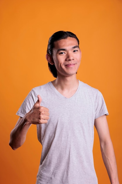 Jeune homme asiatique souriant et joyeux, abandonnant le pouce vers le haut, portrait de plan moyen. Adolescent positif debout avec une expression faciale joyeuse, montrant un geste d'approbation, regardant la caméra