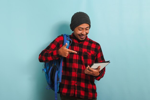 Un jeune homme asiatique souriant pointe vers un livre dans sa main tout en se tenant sur un fond bleu