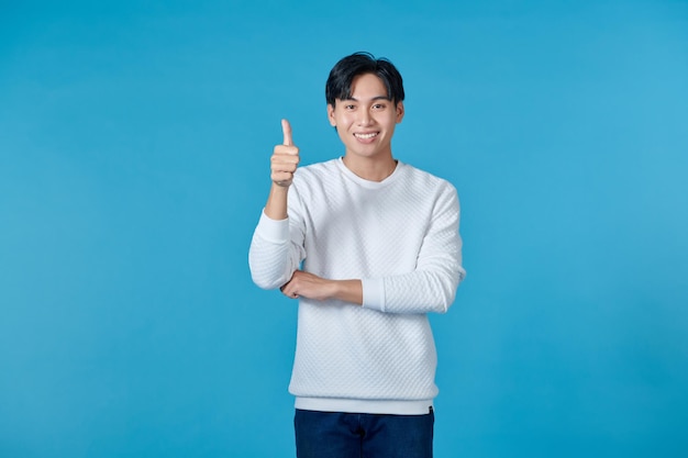 Un jeune homme asiatique souriant portant un pull blanc et un jean sur un fond bleu donne le pouce en l'air