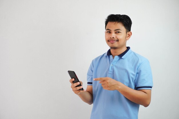 jeune homme asiatique souriant portant un t-shirt bleu tenant un téléphone et pointant vers un smartphone avec le doigt