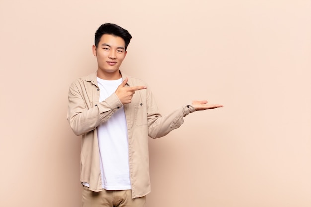 Jeune homme asiatique souriant, se sentant heureux, insouciant et satisfait, pointant vers le concept ou l'idée sur le côté sur le mur de couleur