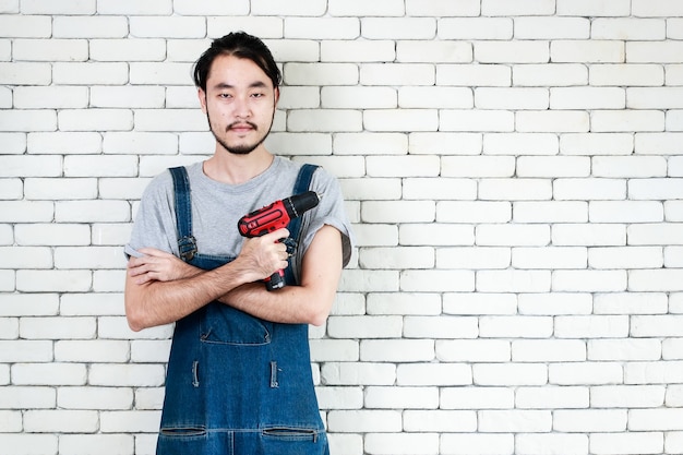 Jeune homme asiatique tenant une perceuse électrique debout devant un mur de briques blanches, souriant et regardant