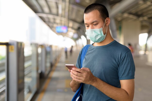 Jeune homme asiatique utilisant un téléphone avec un masque pour se protéger contre l'épidémie de coronavirus à la gare du ciel