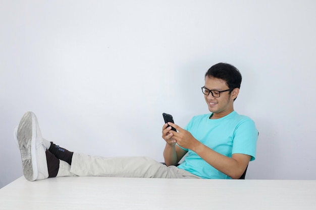 Jeune homme asiatique avec un visage heureux avec ce qu'il voit sur un téléphone portable avec la jambe sur la table Homme indonésien portant une chemise bleue