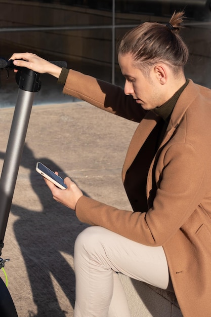 Jeune homme assis activant l'application de scooter électrique sur le smartphone pour le parcourir dans le centre-ville