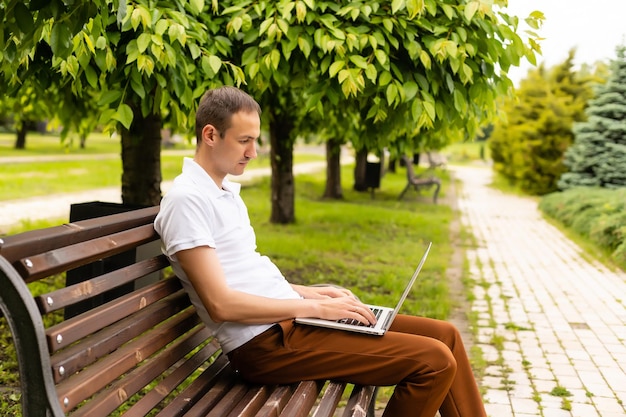 Jeune homme assis sur le banc du parc avec un ordinateur portable sur ses genoux.