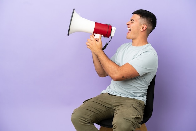 Jeune homme assis sur une chaise sur fond isolé criant à travers un mégaphone