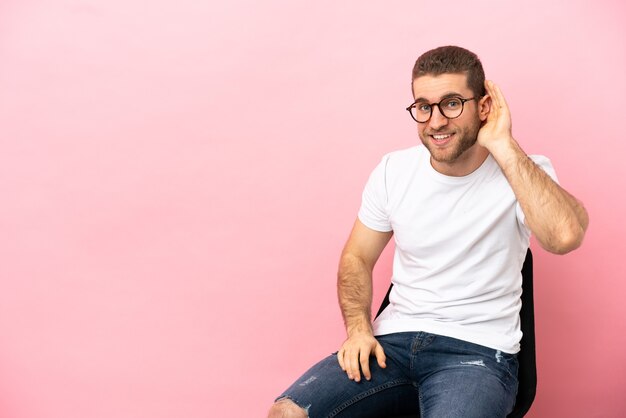 Jeune homme assis sur une chaise sur fond rose isolé en écoutant quelque chose en mettant la main sur l'oreille