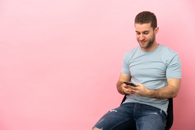 Jeune homme assis sur une chaise sur fond rose isolé envoyant un message avec le mobile