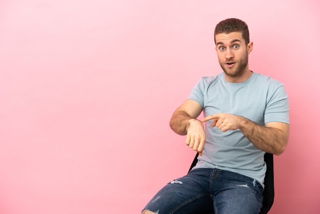 Jeune homme assis sur une chaise sur fond rose isolé faisant le geste d'être en retard