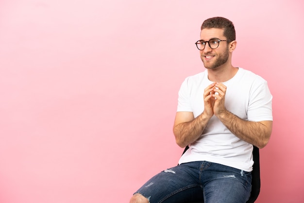 Jeune homme assis sur une chaise sur fond rose isolé intrigant quelque chose