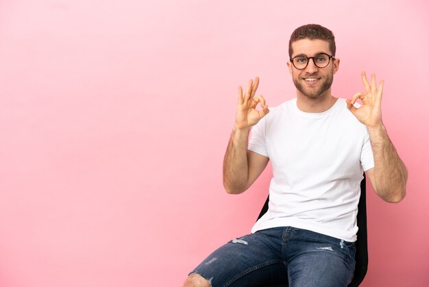 Jeune homme assis sur une chaise sur fond rose isolé montrant signe ok avec deux mains