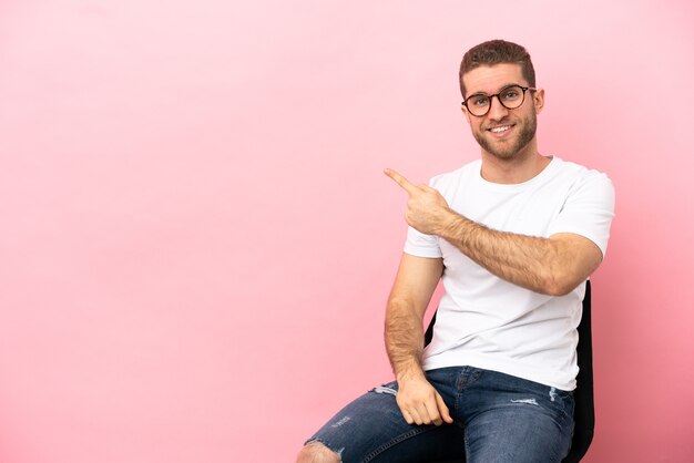 Jeune homme assis sur une chaise sur fond rose isolé pointant vers le côté pour présenter un produit