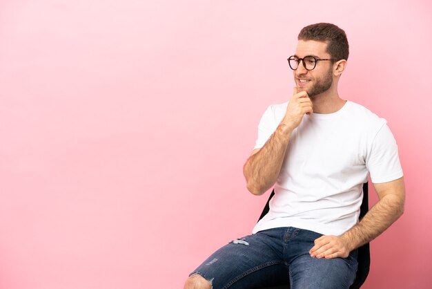 Jeune homme assis sur une chaise sur fond rose isolé à la recherche sur le côté et souriant