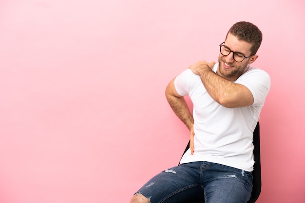 Jeune homme assis sur une chaise sur fond rose isolé souffrant de douleurs à l'épaule pour avoir fait un effort